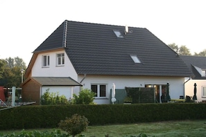A two-story house with a sloped roof, a small attached garage, and a well-maintained front yard with a hedge and a lawn. - Holiday home with 1 bedroom (Zinnowitz)