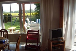 A room with a TV, a chair, a table, and a view of a garden through a window. - Holiday home with 3 bedrooms (Otterndorf)