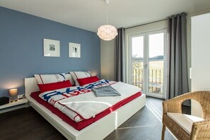 Ein modernes Schlafzimmer mit einem großen Bett, Blick ins Freie durch eine Schiebetür und einem Korbstuhl.