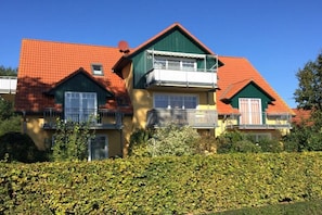 Ein mehrgeschossiges Wohngebäude mit rotem Ziegeldach, Balkonen und einer gepflegten Hecke davor.