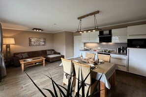 Eine moderne Küche mit weißen Schränken, ein Essbereich mit Korbstühlen und ein Wohnzimmer mit einer Couch und einem Couchtisch.