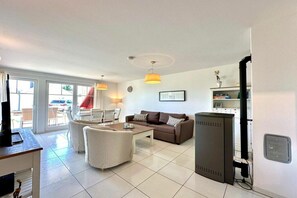 A living room with a sofa, a coffee table, and a television. There is a dining area with a table and chairs. The room has a large window and a door leading outside. - Holiday apartment with 1 bedroom (Sierksdorf)