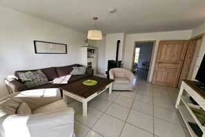 Een woonkamer met een bank, fauteuil, salontafel en een televisie. Er is een open haard en een deur die naar een andere kamer leidt.
