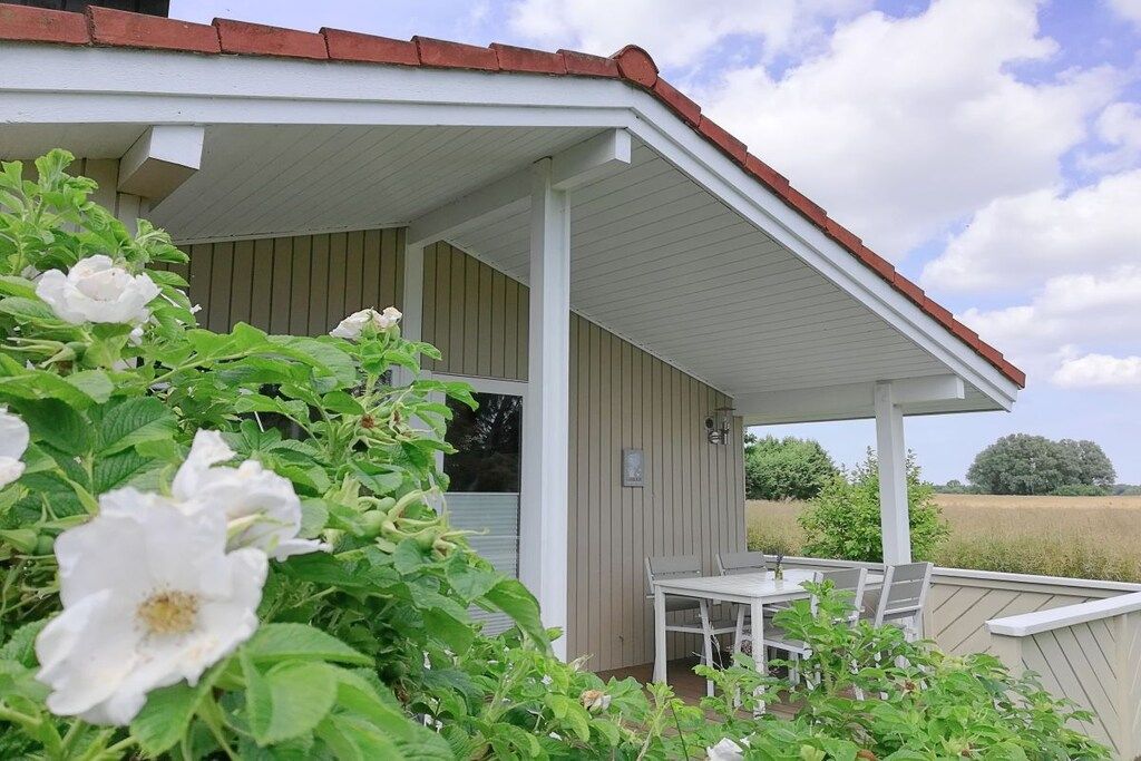 A covered outdoor area with a table and chairs, surrounded by greenery and a building with a sloped roof.