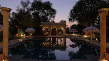A serene pool area with a reflection, surrounded by columns and umbrellas, and a building with arches in the background.