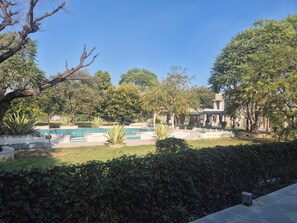 Piscina com deck revestido, cercada por vegetação e um edifício com colunas.