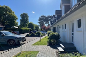 A residential street with parked cars, a white building with a porch, and a clear sky. - Holiday apartment with 1 bedroom (Hohwacht)