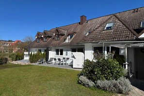 A two-story house with a sloped roof, a covered patio with outdoor furniture, and a well-maintained lawn. - Holiday apartment with 1 bedroom (Hohwacht)