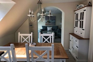 A dining area with a wooden table and white chairs, a chandelier, and a white cabinet with drawers. - Holiday apartment with 1 bedroom (Hohwacht)