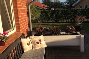 Eine hölzerne Bank mit Kissen, eine Ziegelwand und ein Garten mit verschiedenen Pflanzen und einer Hecke.