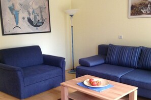 A living room with two blue sofas, a wooden coffee table with apples, a floor lamp, and abstract art on the walls. - Holiday apartment in Söll with swimming pool (Söll)