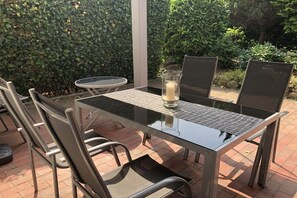 An outdoor patio with a glass-top table and metal chairs, surrounded by a hedge and a brick floor. - on the inland lake in the town center (Heiligenhafen)