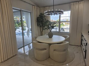 A modern dining area with a round table, cushioned chairs, a vase with greenery, and a view of a pool and palm trees through large windows. - Royal Waterfront 5BR · Private Dock · Sleeps 16 (Port Charlotte)