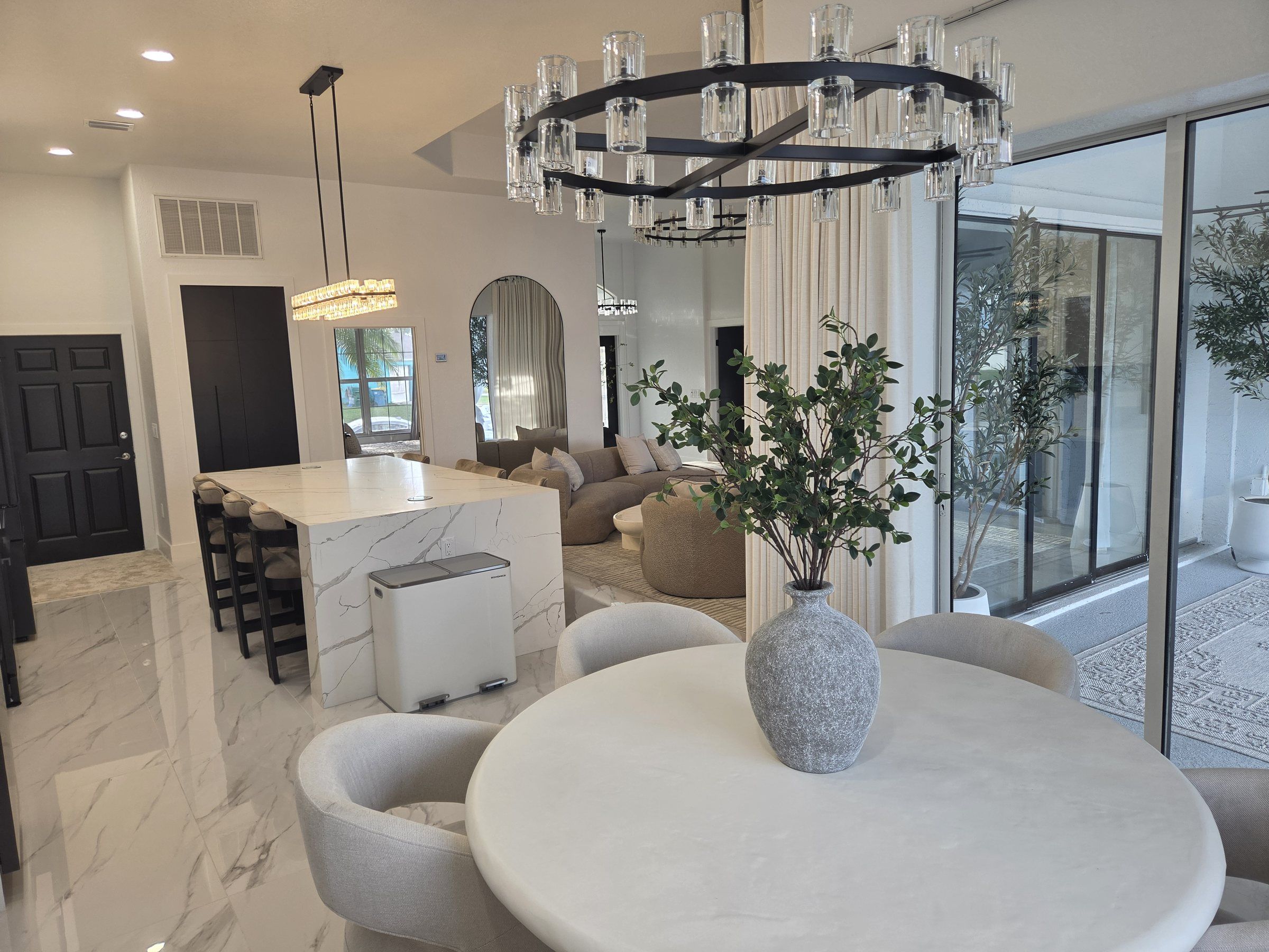 A modern interior with a marble dining table, a central island with bar stools, a chandelier, and a large window with a view.