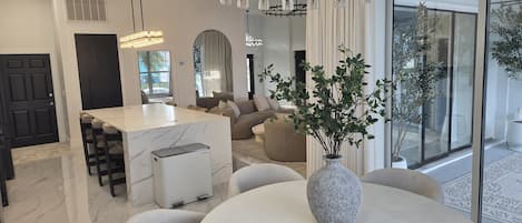 Un intérieur moderne avec une table à manger en marbre, un îlot central doté de tabourets de bar, un lustre et une grande fenêtre offrant une vue imprenable.