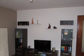 A living room with a flat-screen TV, shelves with decorative items, and a door. - Holiday apartment with 2 bedrooms (Grünhain-Beierfeld)