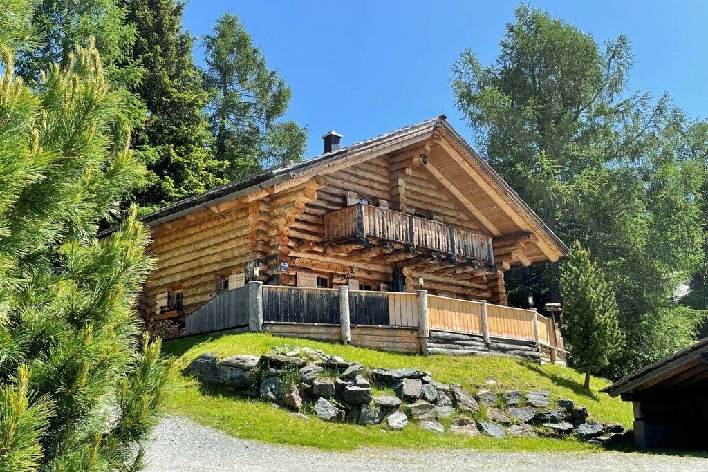 Une cabane en rondins dotée d’un balcon, entourée d’arbres et d’un mur de pierres.