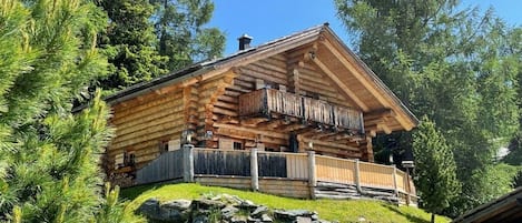 A log cabin with a balcony, surrounded by trees and a stone wall.