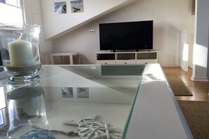 A modern living room with a glass-top coffee table, a flat-screen TV on a white stand, and a decorative glass vase with a candle. - Holiday home with 1 bedroom (Heiligenhafen)
