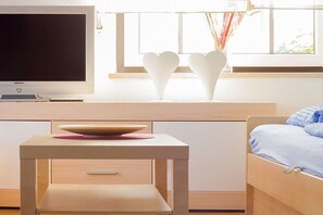 A modern bedroom with a bed, a bedside table with a bowl, a flat-screen TV, and heart-shaped vases on a windowsill. - Holiday home with 1 bedroom (Heiligenhafen)