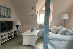 A modern living room with a television, white furniture, a chandelier, and a painting of sailboats. - Holiday apartment with 2 bedrooms (Hohwacht)