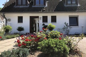 Un bâtiment blanc avec un toit recouvert de tuiles, une porte et des fenêtres. Un jardin avec différentes plantes et fleurs se trouve devant.