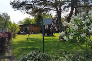 A wooden cabin with a metal roof, a ladder, and a wheelbarrow in a grassy yard with trees and flowering plants. - Summer log cabin "An de Möhl" (Ostseebad Dierhagen \/OT Dändorf)