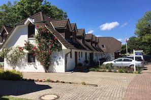 A row of white buildings with brown roofs, a sign indicating a Columbus hotel, and a paved driveway with a car parked nearby. - Holiday apartment with 1 bedroom (Hohwacht)