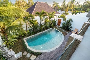 A swimming pool with a curved edge, surrounded by a wooden deck and stone steps leading to a white building with a tiled roof. - Frankie's Villas (Klungkung)