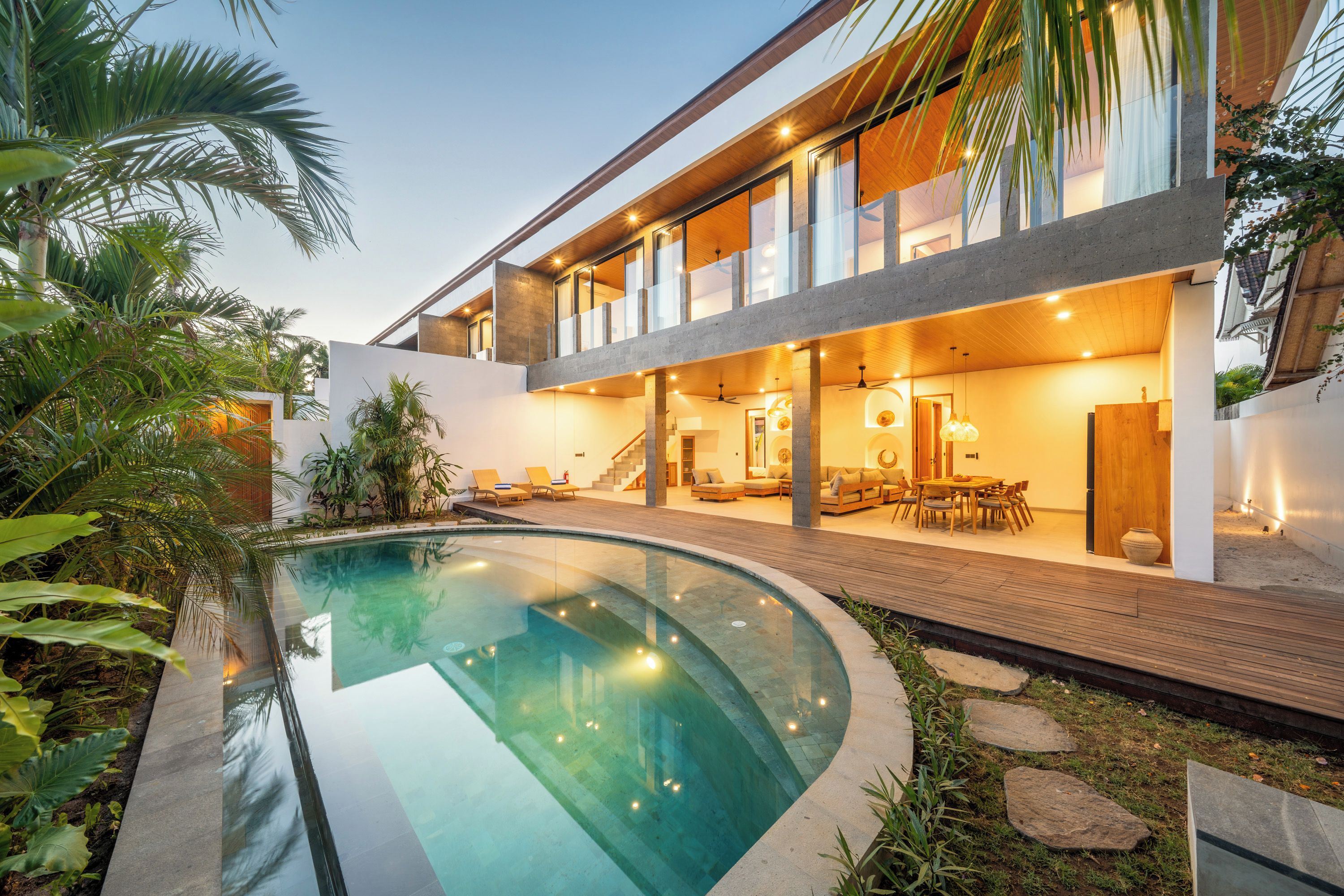 Una casa moderna con piscina curva, terraza de madera y zona de comedor con mobiliario exterior.