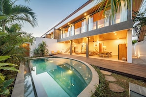 Ein modernes Haus mit einem bogenförmigen Schwimmteich, einer Holzterrasse und einem Essbereich mit Gartenmöbeln.