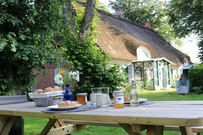 Een picknicktafel met eten en drinken, een broodmand en een glazen pot, buiten geplaatst met een rieten dakgebouw op de achtergrond.