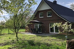 A brick house with a sloped roof, a patio with outdoor furniture, and a tree in the foreground. - Kate on the hiking trail (Drochtersen)