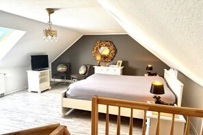 Ein Schlafzimmer mit schrägverdeckter Decke, einem Bett, einem Schminktisch, einem Sessel, einem Fernseher und einer Kronleuchter.