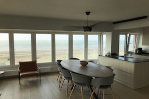 A modern kitchen with large windows overlooking a beach, a dining area with a round table and chairs, and a single chair in the foreground. - Holiday apartment with 1 bedroom (De Haan)