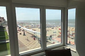 A room with large windows overlooking a beach area with a checkered pavement, a fence, and beachgoers. - Holiday apartment with 1 bedroom (De Haan)