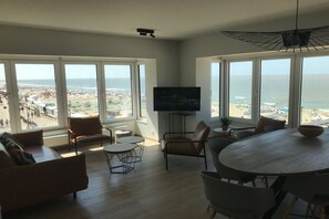 A living room with a large TV, seating, and a view of the beach. - Holiday apartment with 1 bedroom (De Haan)