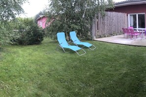 A backyard with a lawn, two blue lounge chairs, a wooden fence, and a pink building in the background. - Holiday home with 2 bedrooms (Neuendorf)