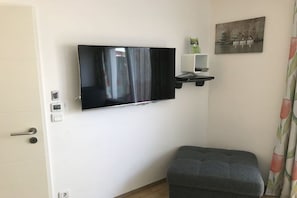 A room with a mounted television, a small shelf with decorative items, a door with a handle, and a patterned curtain. - Holiday home with 2 bedrooms (Neuendorf)