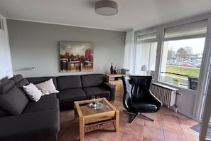 A living room with a sofa, a coffee table, a chair, a painting on the wall, and a view through a window. - Villa by the sea Apt. 39 (Grömitz)