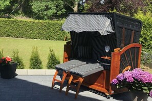 Ein Sitzbereich im Freien aus Rattangeflecht mit gepolsterten Bänken und einem Tisch, umgeben von Grünpflanzen und Blumen.