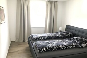 Ein Schlafzimmer mit einem dunkelgrauen Schlafsofa, zwei Kissen mit einem weißen abstrakten Muster, einer Nachttischlampe und einem Fenster mit Vorhängen.