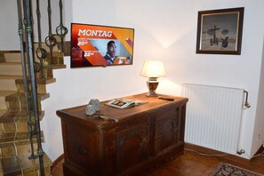 Un escalier orné d’une rampe décorative, une armoire en bois avec une lampe, un tableau encadré accroché au mur et un téléviseur diffusant un événement sportif.