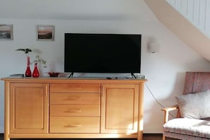 A living room with a wooden cabinet, a flat-screen TV, a chair, and two red vases with plants. - Holiday apartment with 2 bedrooms (Steinberg)