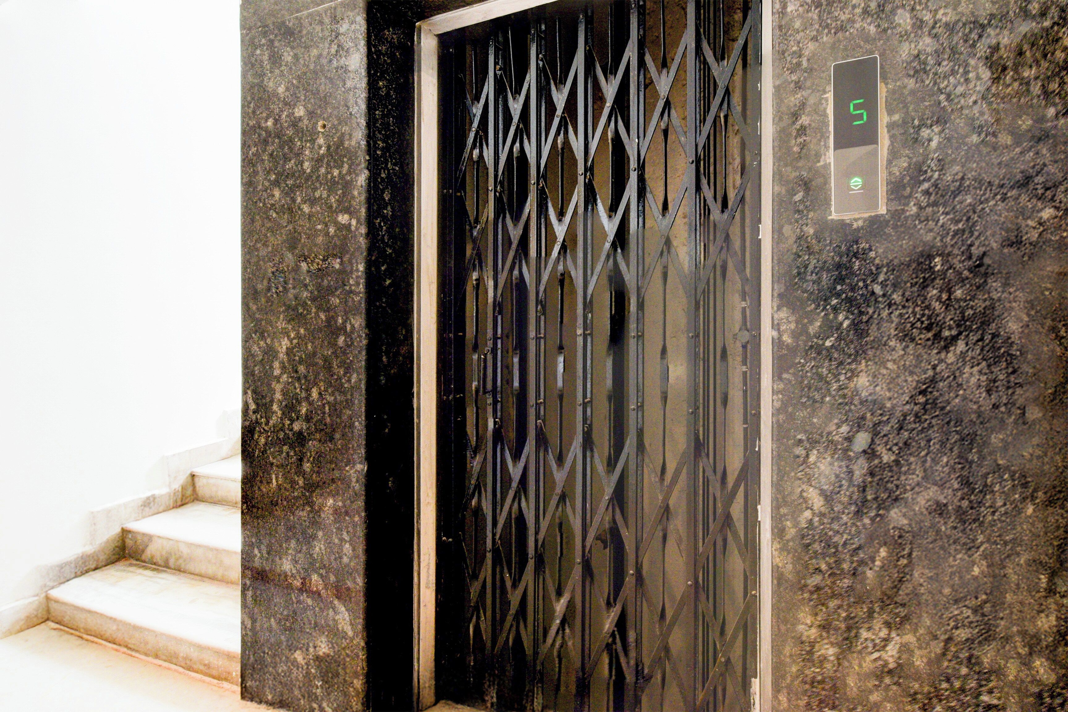 Uma porta de metal preta com padrão geométrico, um painel de controle de elevador digital com o número 5 e uma parede de mármore.