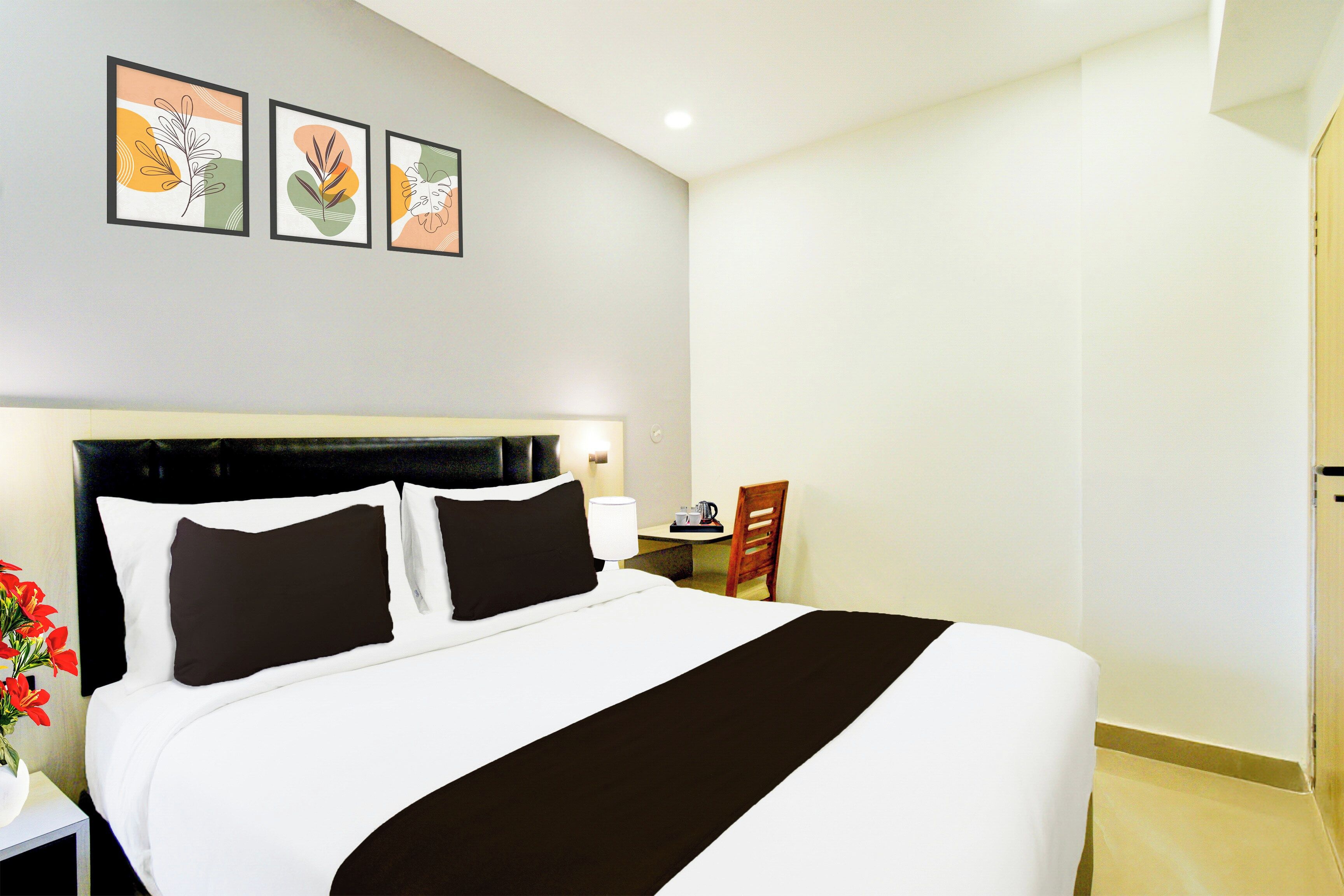 A hotel room with a large bed, a bedside table with a lamp, a chair, and a small table with a cup and a kettle. There are three framed pictures on the wall above the bed.