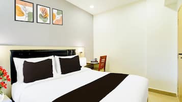A hotel room with a large bed, a bedside table with a lamp, a chair, and a small table with a cup and a kettle. There are three framed pictures on the wall above the bed.