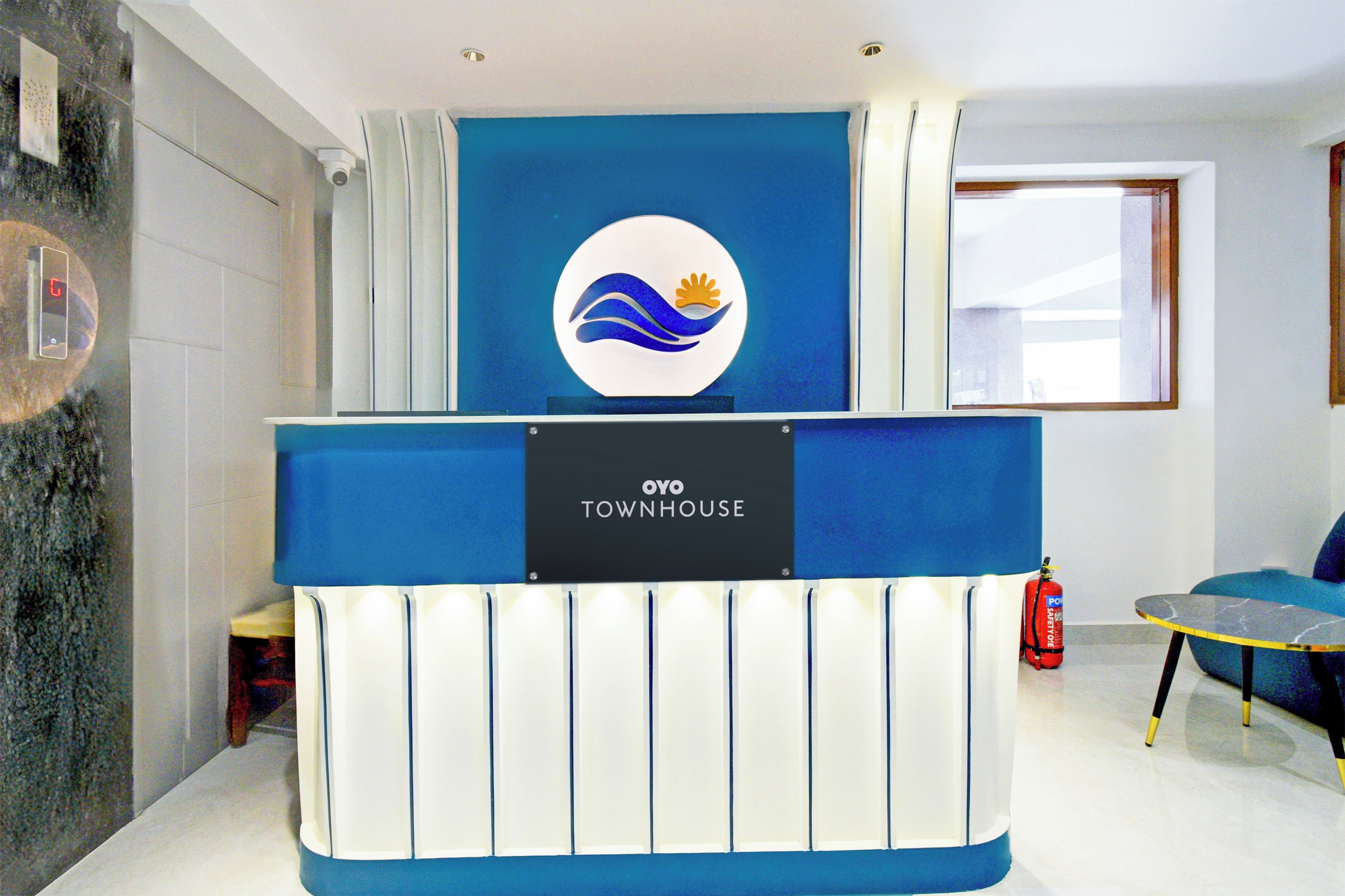 Un mostrador de recepción con logotipo y señalización que indica 'OYO TOWNHOUSE', un extintor y una ventana con persianas.