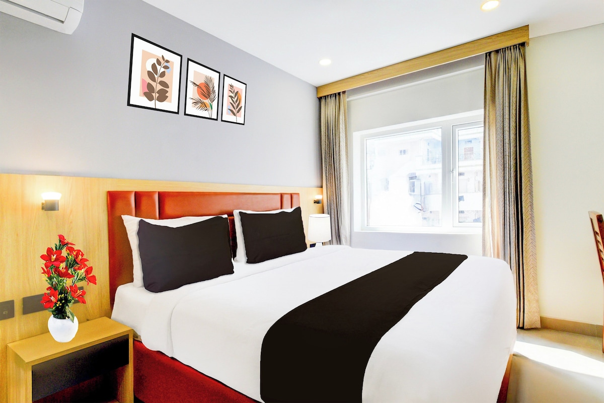 A hotel room with a bed, a nightstand, a vase with flowers, and a window with curtains.
