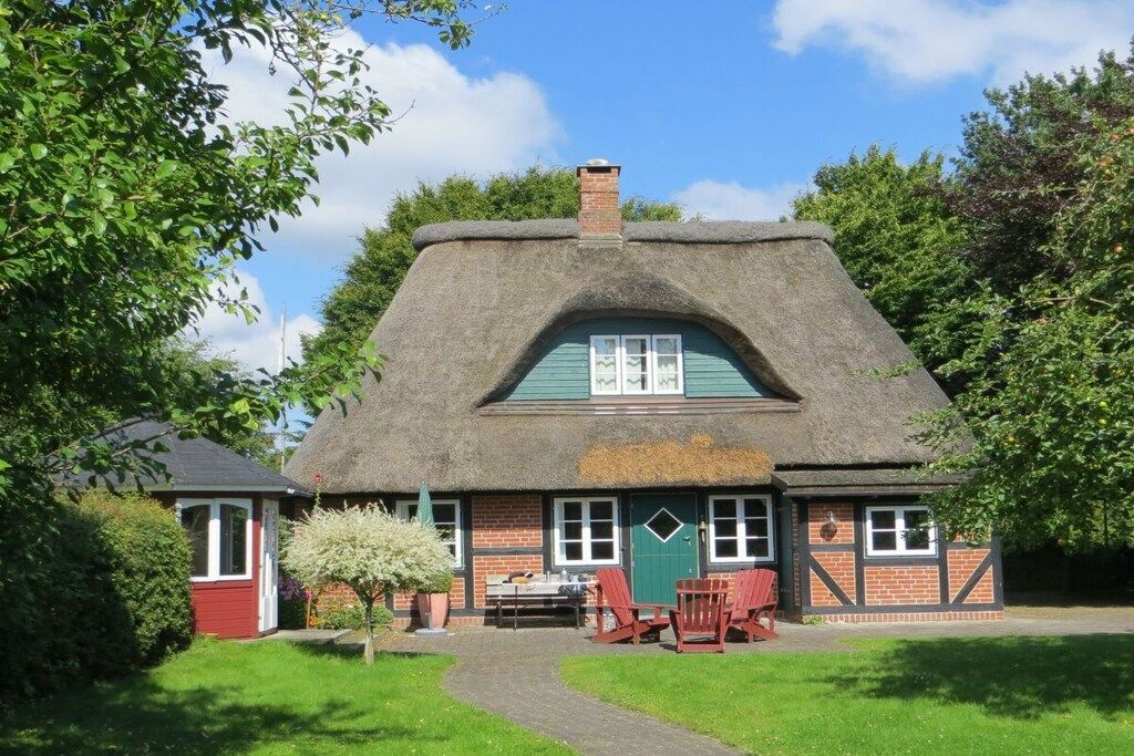 Une maison traditionnelle à toit de chaume, dotée d’une façade en briques, d’un petit jardin et d’un chemin menant à l’entrée.